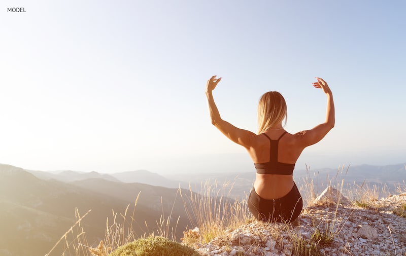 Women sitting on a cliff with her arms lifted, showing her back.