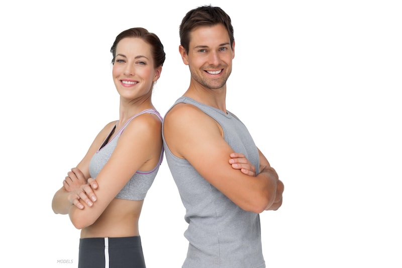 Toned man and woman in workout attire, standing back to back with their arms crossed and smiling at the camera.