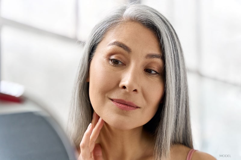 Older woman with gray hair looking, examining her face in a mirror.